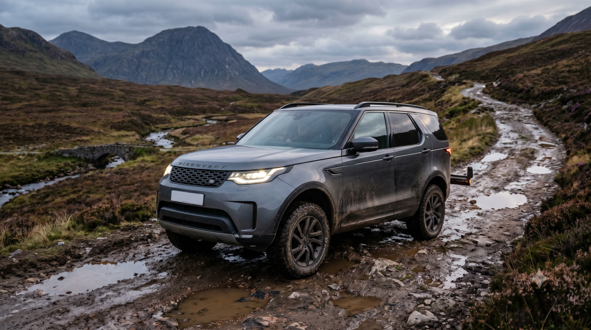land-rover-discovery Land Rover Discovery SUV gris anthracite roulant sur un chemin de terre boueux en montagne sous un ciel couvert